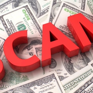 May include: The word SCAM rendered in bold, three-dimensional red capital letters positioned over a scattered collection of United States one hundred dollar bills featuring the portrait of Benjamin Franklin and green security ink details.