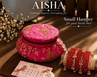 May include: A round, hot pink fabric jewelry box with gold floral embroidery, a set of red crystal bangles with gold charms, and a card featuring the text The House Of AISHA, Small Hamper for your loved once, and a sheet of adhesive bindi stickers.