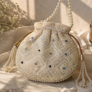 May include: A cream-colored drawstring pouch bag featuring intricate embroidery, sequins, and small white pearls. The bag is adorned with scattered blue crystals and includes a pearl-beaded handle and two side tassels made of hanging pearl strands.
