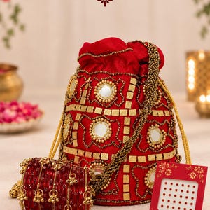 May include: A red velvet drawstring potli bag decorated with gold beads, square metallic accents, and circular mirrors. Beside it are red glass bangles with gold charms and a red card labeled ABR Bindi with small red adhesive dots.