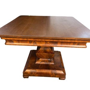 May include: A square wooden table with a dark brown flat top and a thick, pedestal-style base. The base features a prominent, curved, and flared design with a rich, polished wood grain pattern that extends to the square foundation.