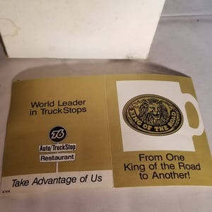 May include: A gold and white advertisement for a truck stop chain called "76 Auto/TruckStop Restaurant". The ad features a lion logo with the text "King of the Road" and the slogan "From One King of the Road to Another!"
