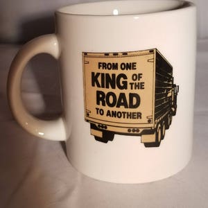 May include: White ceramic coffee mug with a black and brown illustration of a semi-truck. The text on the truck reads "From one King of the Road to another."