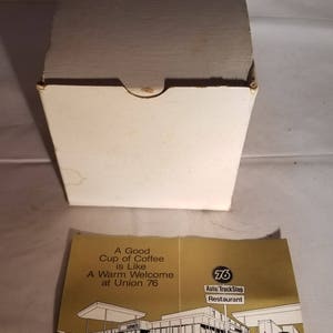 May include: A white box with a gold and white label that reads "A Good Cup of Coffee is Like A Warm Welcome at Union 76". The label also features a drawing of a gas station with the text "76 Auto/TruckStop Restaurant" and "Ready to Serve! Truck Industry Sales. Union Oil Company of California".