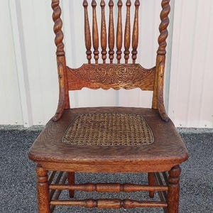 May include: Antique wooden chair with a woven seat. The chair features a carved back with decorative details and turned spindles. The legs and frame are made of dark wood, showcasing the natural grain. A classic piece of furniture.