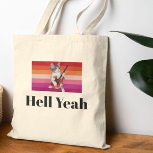 May include: A cream-coloured canvas tote bag featuring a graphic of a cat playing an electric guitar against a horizontal striped background of orange, pink, and red. Below the graphic, the text "Hell Yeah" is printed in bold black letters.