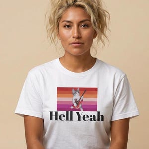 Könnte beinhalten: Ein weißes kurzärmeliges T-Shirt mit einer Grafik einer kleinen Katze, die eine rote E-Gitarre spielt, zentriert über einem horizontal gestreiften Hintergrund in Orange-, Rosa- und Rottönen, mit dem Text Hell Yeah in Schwarz unter der Grafik.