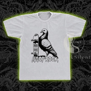 May include: A light gray short-sleeve t-shirt featuring a black graphic of a pigeon standing next to a skateboard. The skateboard deck displays the text MUST NOT SLEEP MUST WARN OTHERS, and the name AESOP ROCK is printed below in a dripping font.