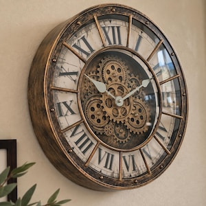 May include: A round wall clock with a distressed bronze-coloured frame and a cream-coloured face featuring black Roman numerals. The centre of the clock displays an exposed mechanism of interconnected gold-toned gears with two simple white hands.