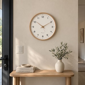 May include: A round wall clock with a light wood frame, a white face, and black Arabic numerals. The clock features thin, light-colored hands and a small logo near the center. The design includes minute markers around the perimeter of the face.