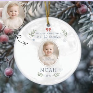 May include: A round white ceramic Christmas ornament featuring a gold hanging string. The design includes a central photo placeholder, a red bow graphic, green leaf accents, and the text: My First Christmas as a big brother, Noah, 2026.