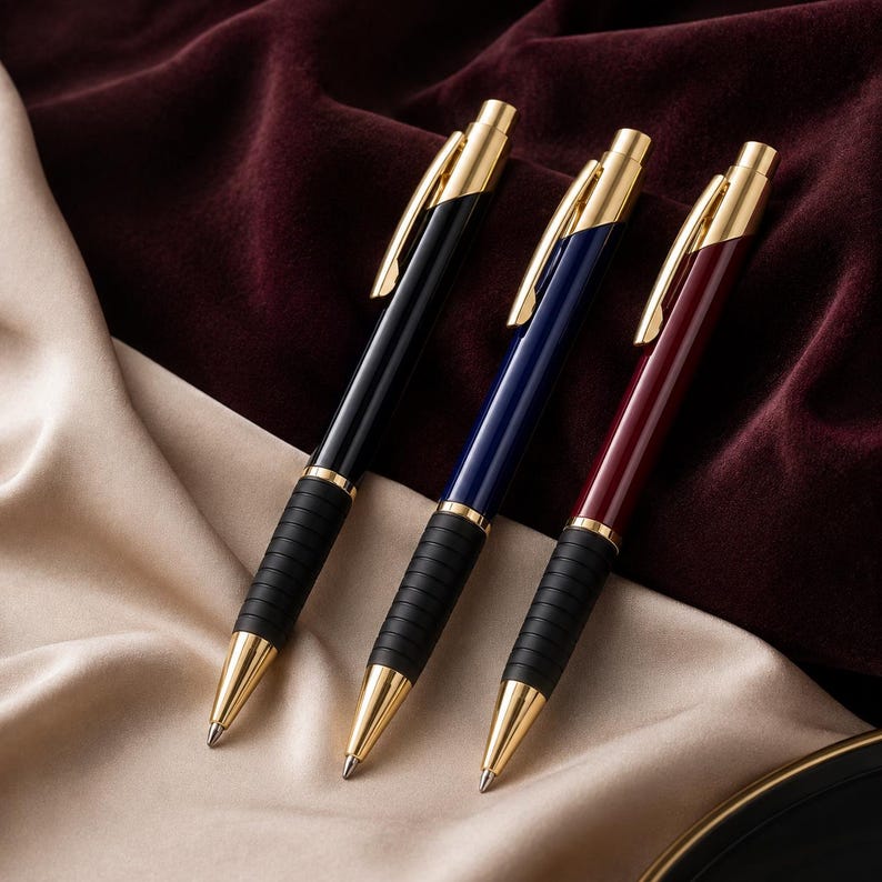 K&ouml;nnte beinhalten: Drei einziehbare Kugelschreiber mit goldfarbenen Clips, Spitzen und Mittelringen. Jeder Stift verf&uuml;gt &uuml;ber eine gerippte schwarze Gummigriffzone und einen gl&auml;nzenden Schaft in Schwarz, Marineblau und Dunkelrot, die diagonal angeordnet sind.
