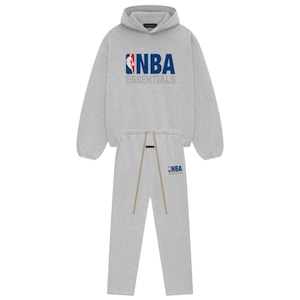 May include: A light gray matching set consisting of a long-sleeve hooded sweatshirt and sweatpants. The hoodie features the NBA logo and the word ESSENTIALS in blue and white. The sweatpants include a small NBA logo and long tan drawstrings.