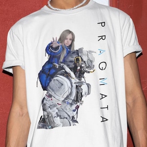 May include: A white short-sleeved t-shirt featuring a graphic print of a young person with long blonde hair wearing a blue jacket, riding on the shoulders of a grey and white robotic figure. The text PRAGMATA is printed vertically in black.