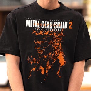 May include: A black short-sleeved t-shirt featuring an orange graphic print of a character and the text "METAL GEAR SOLID 2 SONS OF LIBERTY" in white and orange lettering across the chest, with "TACTICAL ESPIONAGE ACTION" printed above the title.