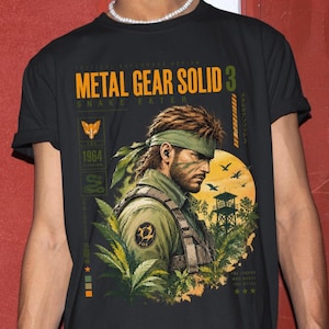 Może przedstawiać: Czarny t-shirt z nadrukiem graficznym przedstawiającym postać Naked Snake z gry Metal Gear Solid 3: Snake Eater. Projekt zawiera żółty napis 'METAL GEAR SOLID 3' i 'SNAKE EATER', dżunglę, emblemat lisa oraz datę 1964.