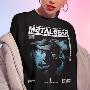 Könnte beinhalten: Ein schwarzes Kurzarm-T-Shirt mit einem Grafikdruck und dem Text "TACTICAL ESPIONAGE ACTION METAL GEAR SOLID". Das Design enthält ein blau getöntes Porträt, ein "99.99" Preisschild, ein Waffensymbol und den Text "PRESS START BUTTON".