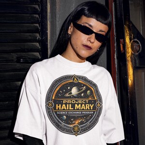 May include: A white short-sleeved t-shirt featuring a circular graphic on the chest. The design depicts a space scene with planets and stars, surrounded by a border with scientific icons. Text reads: PROJECT HAIL MARY SCIENCE EXCHANGE PROGRAM.