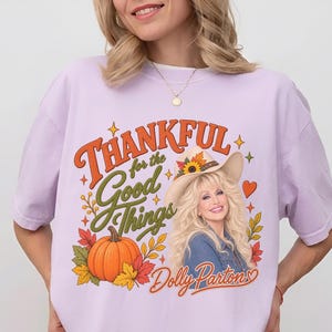 May include: A light purple t-shirt featuring a graphic print with the text Thankful for the Good Things and Dolly Parton. The design includes a portrait of Dolly Parton wearing a cowboy hat, an orange pumpkin, autumn leaves, and small stars.