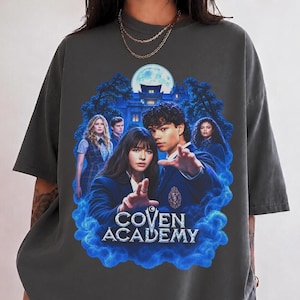 May include: A charcoal grey short-sleeve t-shirt featuring a graphic print of four individuals in school uniforms. The design includes a large building under a full moon, blue smoke effects, and the text COVEN ACADEMY in a bold, stylised font.