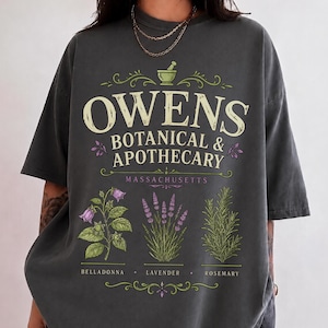 Könnte beinhalten: Ein anthrazitfarbenes T-Shirt mit einem Grafikdruck, der OWENS BOTANICAL & APOTHECARY MASSACHUSETTS zeigt. Das Design enthält ein Mörser- und Stößel-Symbol sowie Illustrationen von Tollkirsche, Lavendel und Rosmarin mit den jeweiligen Namen.