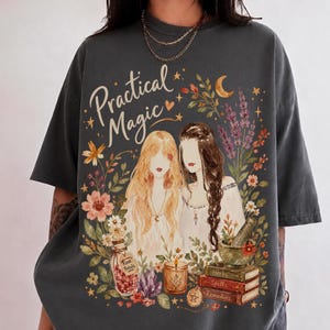 Puede incluir: Una camiseta gris carbón con un estampado gráfico de dos figuras de pelo largo rodeadas de flores, una luna creciente, una botella con la etiqueta Rose Petals, una vela, un mortero y una pila de libros con los títulos Herbs, Spells, Remedies.