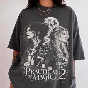 Op de afbeelding: Een antracietgrijs T-shirt met korte mouwen en een grafische print van twee vrouwen, een groot huis, een volle maan, vogels, een zwarte kat, kaarsen en bloemen. De tekst "PRACTICAL MAGIC 2" staat onderaan in een wit schreeflettertype gedrukt.