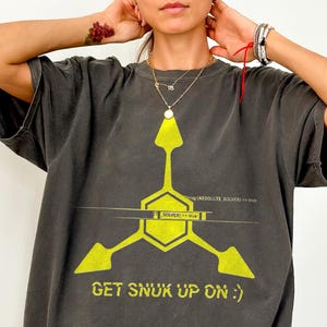 Könnte beinhalten: Ein anthrazitfarbenes Kurzarm-T-Shirt mit einer großen, leuchtend gelben Grafik in Form eines dreizackigen geometrischen Symbols mit einem zentralen Sechseck. Das Design enthält den Text "GET SNUK UP ON :)" und "SOLVER == true".