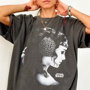 Pode incluir: Uma t-shirt de manga curta em cinzento carvão com uma grande impressão gráfica a preto e branco da Princesa Leia de Star Wars de perfil. O design inclui o logótipo Star Wars em texto branco no lado inferior direito do gráfico.