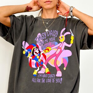 Könnte beinhalten: Ein anthrazitfarbenes Kurzarm-T-Shirt mit einer bunten Grafik eines Hofnarren und einer rosa Hasenfigur, die Spielzeugpistolen halten. Der Text lautet: DAISY DAISY GIVE ME YOUR ANSWER DO! I'M HALF CRAZY ALL FOR THE LOVE OF YOU! in lila Schrift.