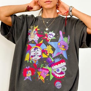 May include: A charcoal gray short-sleeve t-shirt featuring a vibrant graphic print of various colorful cartoon characters and star shapes in shades of purple, pink, yellow, and red. The design includes stylized faces, expressive eyes, and jagged teeth patterns.