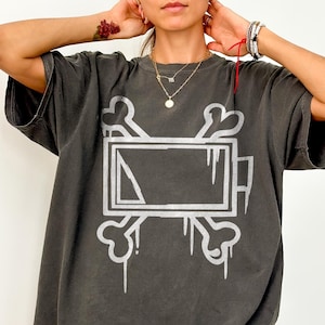 May include: A charcoal grey short-sleeve t-shirt featuring a large, distressed white graphic print on the front. The design depicts a rectangular battery icon centred over two crossed bones, creating a skull and crossbones style motif.