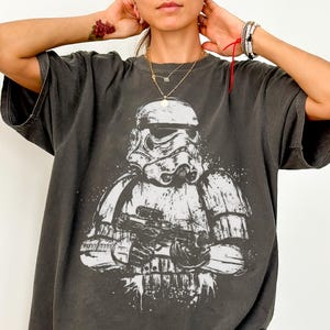May include: A charcoal gray short-sleeve t-shirt featuring a large, distressed white graphic print of a Stormtrooper holding a blaster rifle. The shirt has a relaxed, oversized fit with a crew neckline and a faded, vintage-inspired wash.