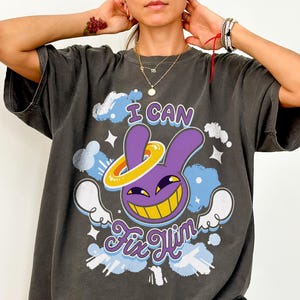 Könnte beinhalten: Ein anthrazitfarbenes Kurzarm-T-Shirt mit einer Grafik eines violetten Hasencharakters mit gelbem Heiligenschein und breitem Grinsen. Der Text "I CAN" steht über der Figur und "Fix Him" darunter, umgeben von hellblauen Wolken und weißen Sternen.
