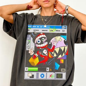 Könnte beinhalten: Ein anthrazitfarbenes T-Shirt mit einem Grafikdruck eines digitalen Schnittstellenfensters. Das Design zeigt eine Figur mit Zylinder und rotem Anzug neben einer runden, zahnigen Kreatur. Die Benutzeroberfläche enthält Menüleisten, Symbole und Text.