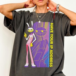 May include: A charcoal gray short-sleeve t-shirt featuring a graphic of a cartoon rabbit character in pink overalls. The design includes a purple and yellow halftone background with the text HAVE YOUR IP ADDRESS printed vertically in yellow.
