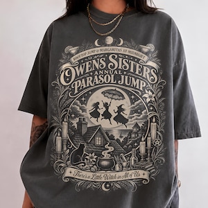 以下が含まれることがあります： 「Owens Sisters Annual Parasol Jump」および「There's a Little Witch in All of Us」というテキストがプリントされたチャコールグレーのTシャツ。デザインには、傘を持ってジャンプする3人のシルエット、家、猫、キャンドルが描かれています。