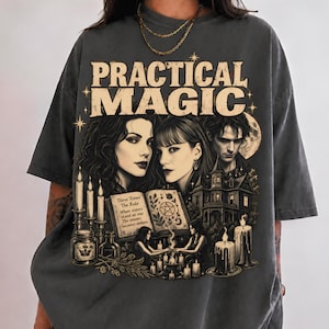 Puede incluir: Una camiseta de manga corta gris carbón con un estampado gráfico que incluye el texto "PRACTICAL MAGIC" en la parte superior. El diseño muestra a dos mujeres, una casa grande, un libro abierto con un pentagrama, velas y estrellas.