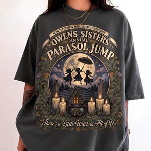 May include: A charcoal grey t-shirt featuring a graphic print of three figures jumping with a parasol under a full moon. The text reads "Owens Sisters Annual Parasol Jump" and "There's a Little Witch in All of Us" above a house, a cauldron, and candles.
