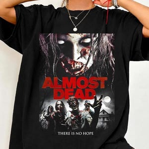 May include: A black short-sleeve t-shirt featuring a graphic print of a zombie movie poster. The design includes the text ALMOST DEAD in large red letters, a close-up of a zombie face, a group of zombies, a house, and the text THERE IS NO HOPE.