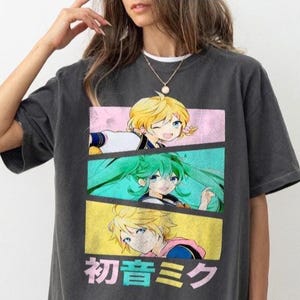 May include: A charcoal gray short-sleeve t-shirt featuring a graphic print of three anime characters in stacked rectangular panels. The design includes a blonde character, a teal-haired character, and a blonde character, with Japanese text below.