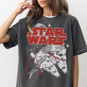 May include: A charcoal grey short-sleeve t-shirt featuring a large red Star Wars logo across the chest. Below the text is a white graphic illustration of the Millennium Falcon spaceship in flight, surrounded by smaller starships and red laser beams.