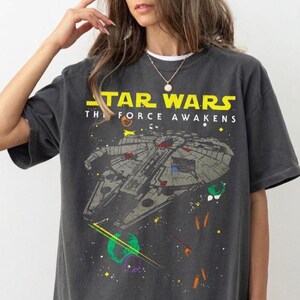 May include: A charcoal grey short-sleeved t-shirt featuring a graphic print of the Millennium Falcon spaceship from Star Wars: The Force Awakens. The design includes yellow text, colourful planets, and star patterns against the dark fabric background.