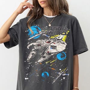 以下が含まれることがあります： スター・ウォーズのミレニアム・ファルコンとXウィング・スターファイターのグラフィックプリントが施された、チャコールグレーの半袖Tシャツ。暗い背景に青い惑星、黄色い光の筋、白い星が描かれています。