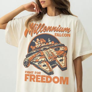 Può includere: Una t-shirt a maniche corte color avorio con una stampa grafica dell'astronave Millennium Falcon. Il design include il testo "Millennium FALCON" e "FIGHT FOR FREEDOM" in arancione e blu scuro su uno sfondo circolare blu scuro.