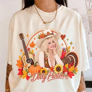 Op de afbeelding: Een crèmekleurig t-shirt met een grafische print van Dolly Parton met een cowboyhoed, omringd door een akoestische gitaar, een pompoen, zonnebloemen, herfstbladeren en een kalkoen. De tekst Dolly Parton staat in rode letters op het shirt.