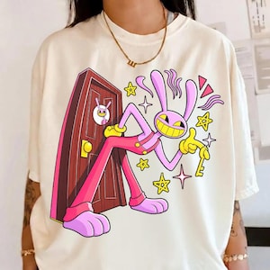 May include: An ivory t-shirt featuring a graphic print of a pink cartoon rabbit with a wide yellow grin, wearing pink overalls, standing next to a brown door. The rabbit holds a yellow key, surrounded by yellow stars and pink abstract shapes.