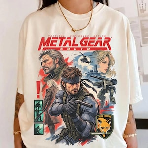 May include: An ivory short-sleeve t-shirt featuring a graphic print from Metal Gear Solid. The design includes characters, a helicopter, a mechanical walker, and the text Metal Gear Solid. A Fox Hound emblem with a fox head is visible at the bottom.