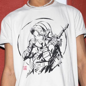 May include: A white short-sleeved t-shirt featuring a black and white ink-style graphic illustration of a character holding an ocarina with a sword strapped to their back, enclosed in a circular brushstroke frame with a small red square stamp.