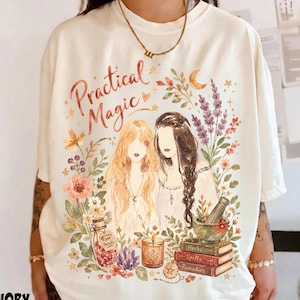 May include: An ivory t-shirt featuring a graphic illustration of two figures with long hair surrounded by flowers, lavender, a bottle labeled Rose Petals, a candle, and a stack of books labeled Herbs, Spells, and Remedies with the text Practical Magic.
